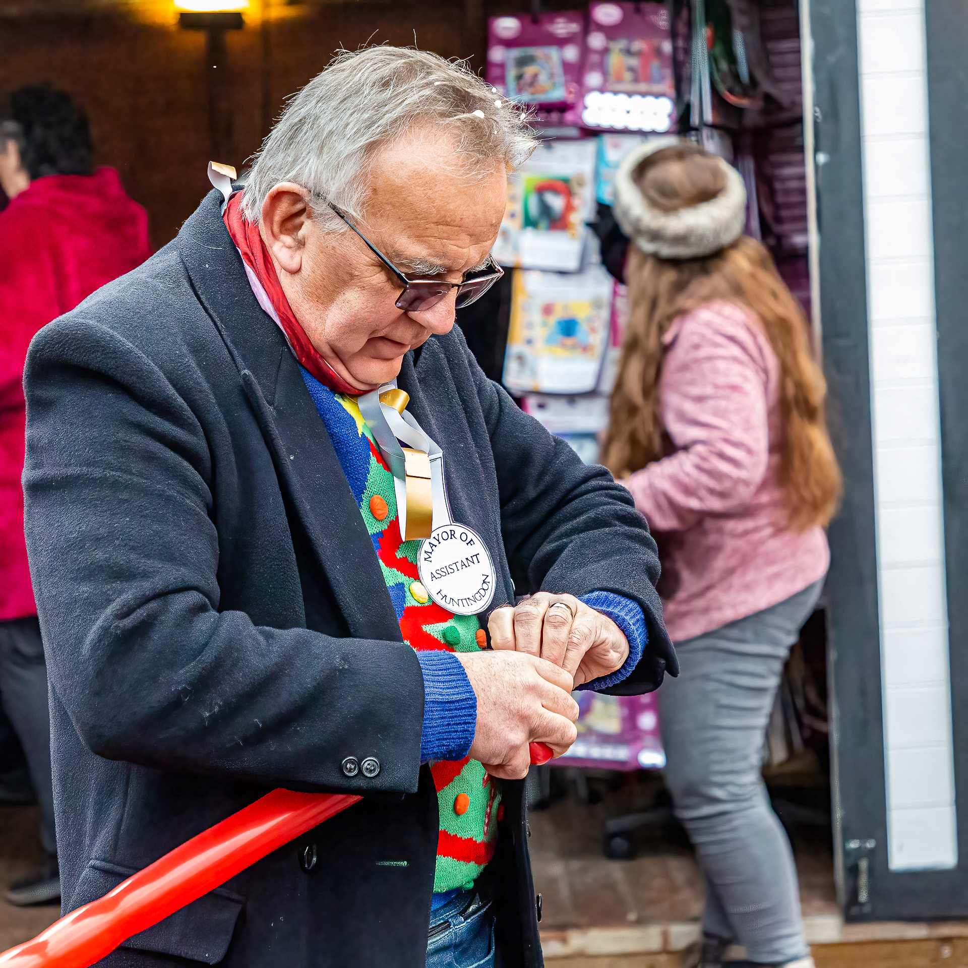 Dave the Balloon man wearing a smart jackets and a bright colour Christmas jumper tying off a long red balloon.