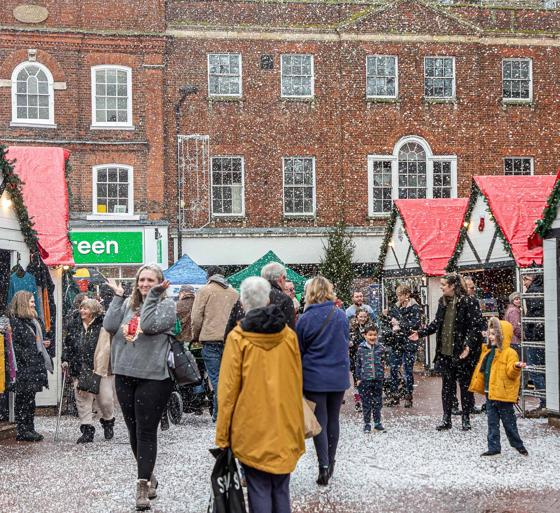 A crowd of shoppers on Huntingdon Market Square, walking between Christmas wooden chalets with red roofs. Gentle snow falls and some children are playing in the foam.
