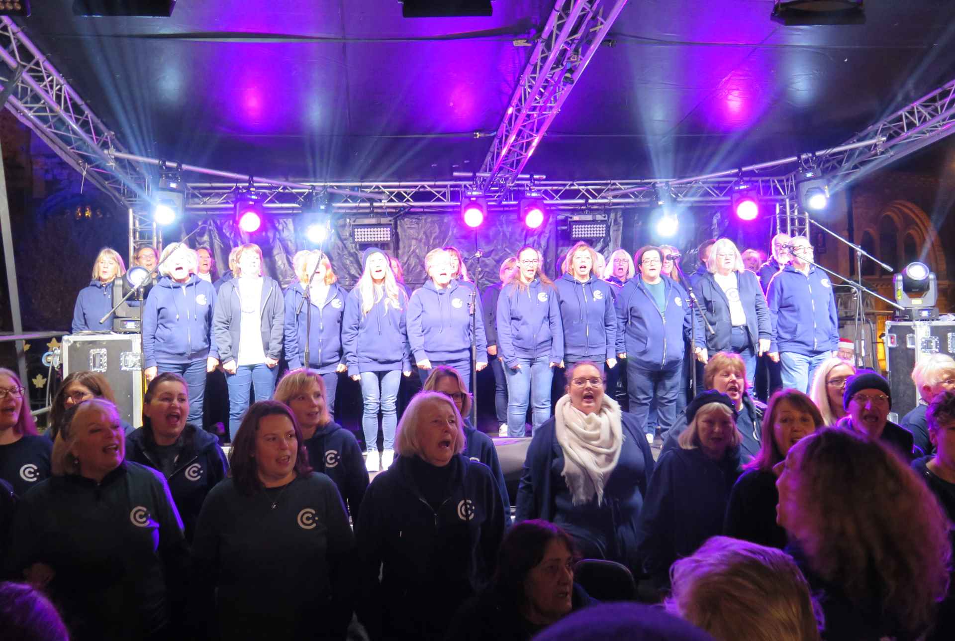 A picture of the Collaboration Choir on stage at Huntingdon Christmas Market. They are all singing passionately and brightly lit with blue an purple lights.  A large crowd watches them perform.