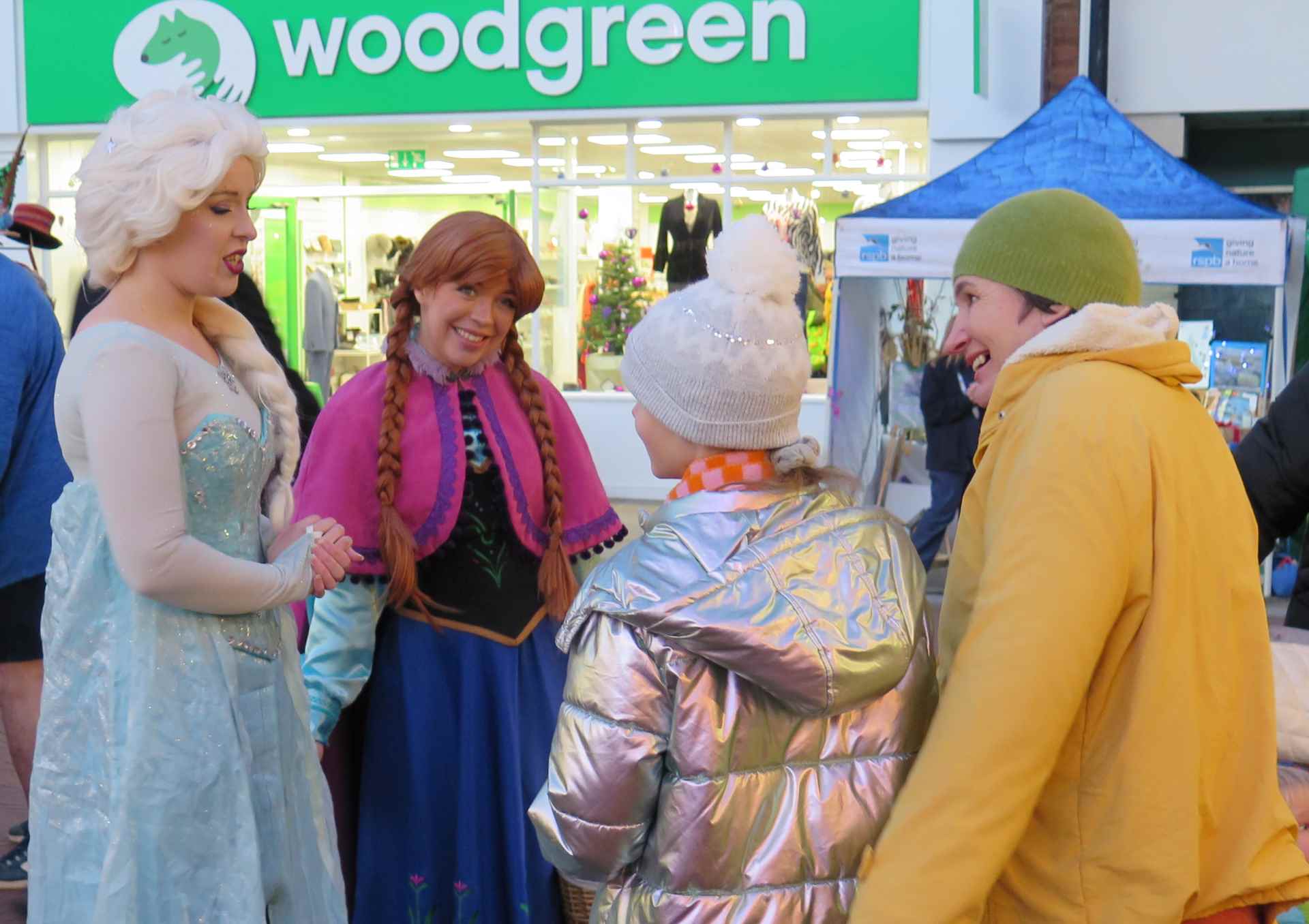 The Ice Princesses pictured talking to a family. One princess is dressed in a light blue flowing glittering dress and with a long white plaited braid. The second princess has two ginger plaits with a pink and blue dress.