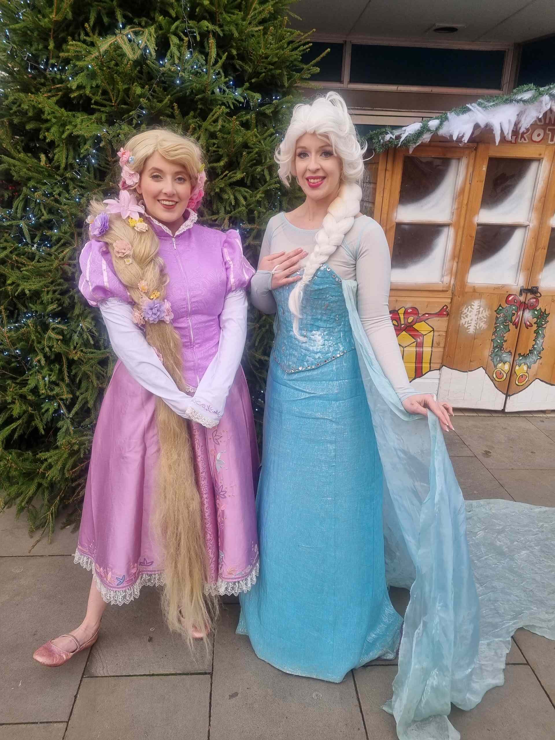 Two princesses pose in front of a Christmas tree and a wooden elf workshop shed. The first princesses has on a floor length pink dress with capped sleeves and long braided hair down to her feet with flowers embedded in her hair. The second Ice princess has a floor length light blue dress that glitters, with white sleeves and a sheer cape that fans out on the floor behind her.