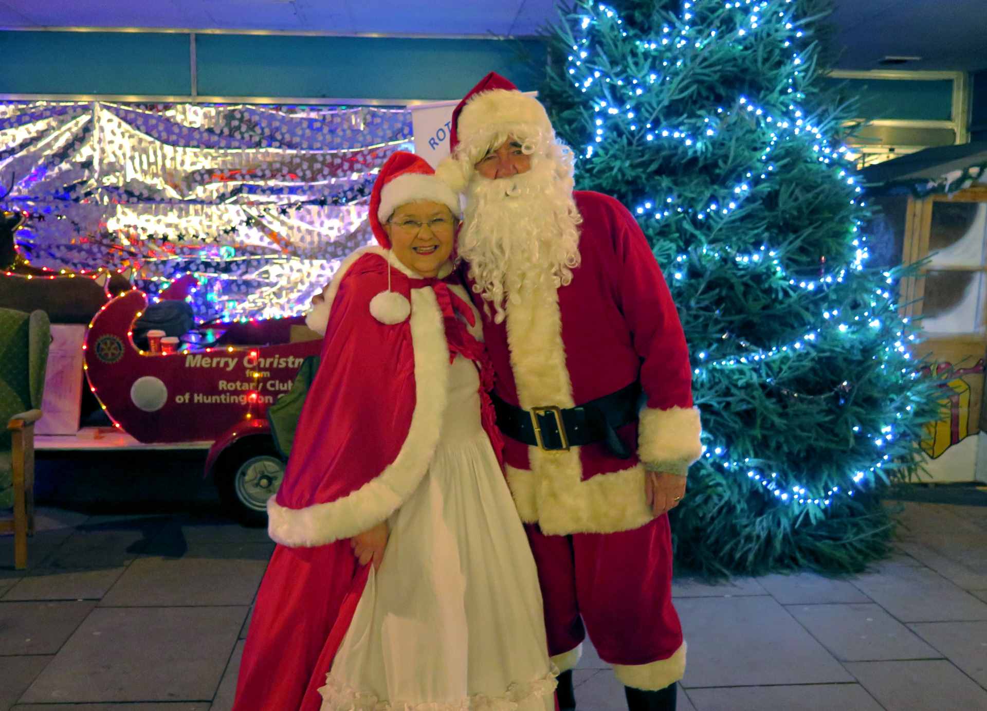 Mrs Claus wearing a white dress with a red and white trimmed shawl and Christmas hat, posing with Father Christmas, in his red suit and white beard, in front of a wooden red Santa's sleigh and Christmas tree illuminated with blue Christmas lights.