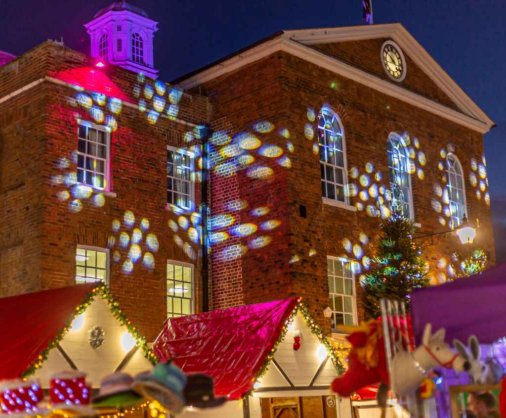Huntingdon Town Hall at night time, images of white snowflakes are projected onto the building. Christmas wooden chalets with red roofs and festoon lighting sit in front of the building.